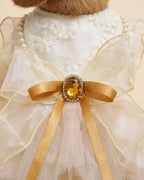 Macro close-up of the champagne organza bow with an amber-colored jewel centerpiece and gold satin ribbon tails on floral lace fabric.