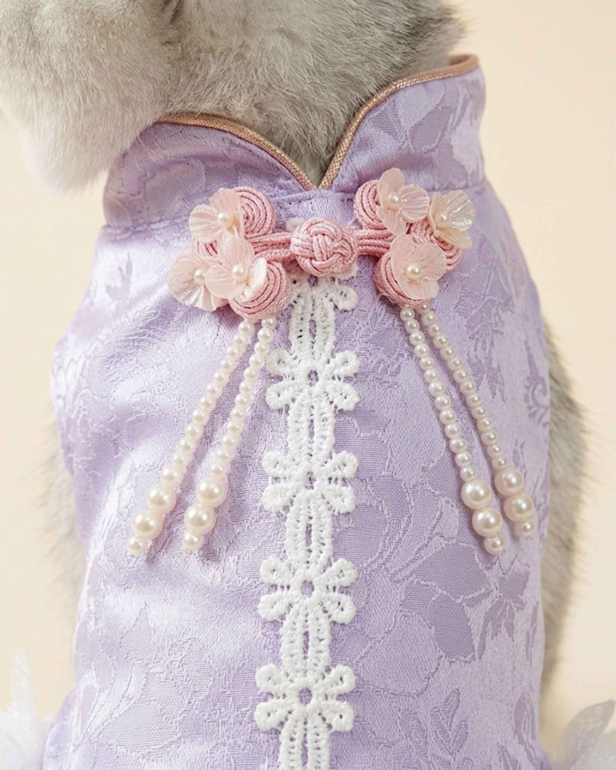 Close-up of pink floral knot ornaments, hanging pearl tassels and white lace trim on a lavender jacquard pet dress.